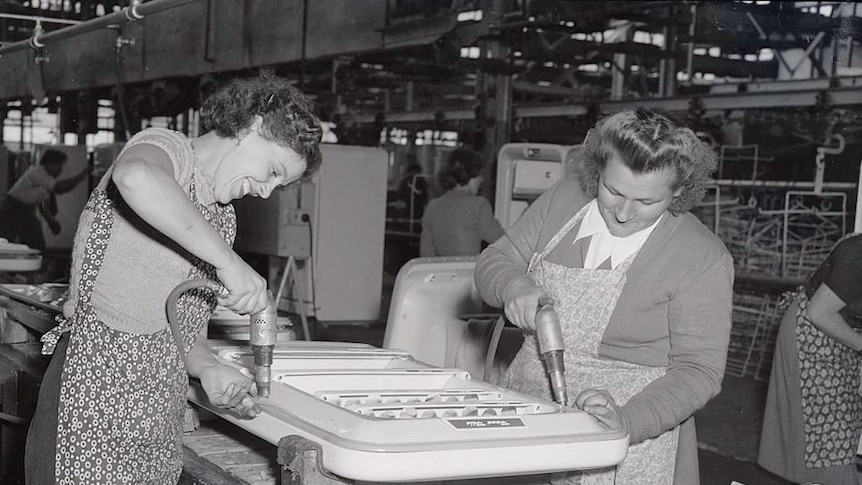 A black and white photo of two women breaking through refrigerator doors at the Emmco factory in 1956.